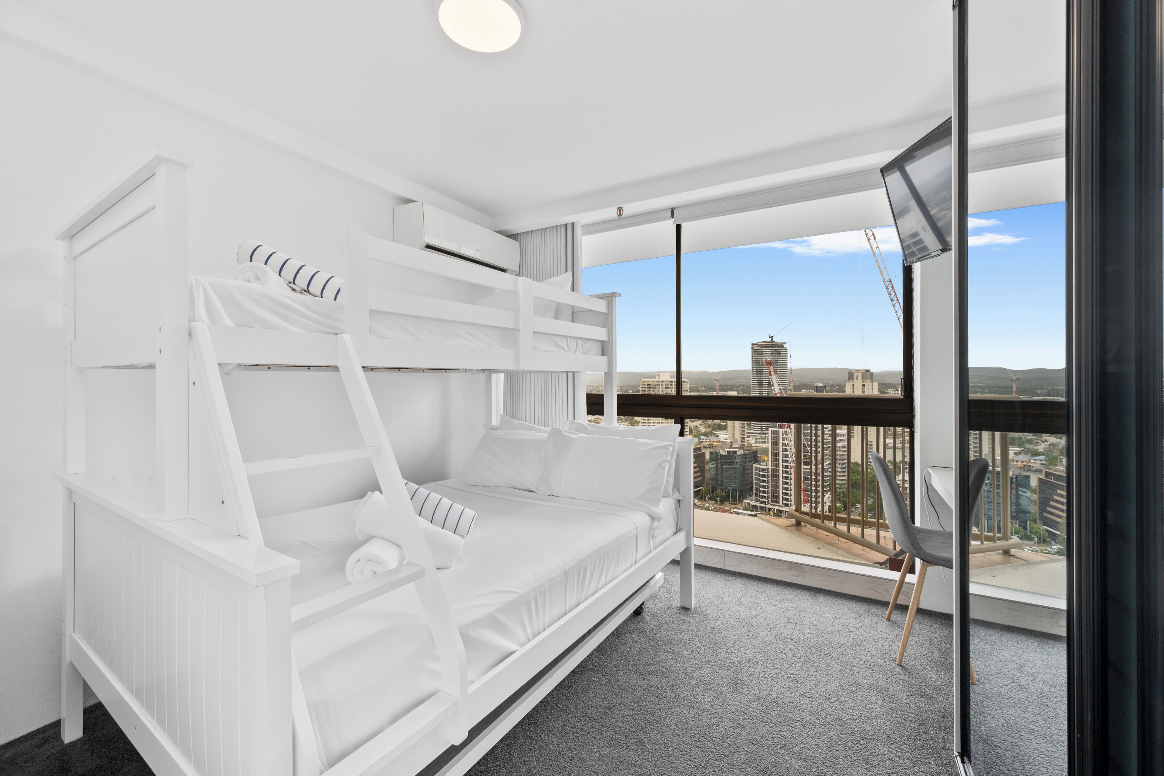 A white bunk bed with a city view, a small table, and a chair in a modern room with large windows.