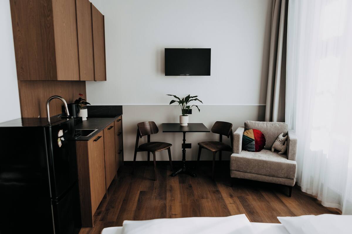 A compact kitchen with a black refrigerator, wooden cabinets, a dining table with two chairs, a small sofa with pillows, and a TV mounted on the wall.