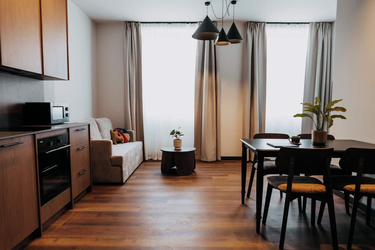 A modern kitchen with wooden cabinets, a built-in oven, and a microwave. There is a dining area with a table, chairs, and a potted plant.
