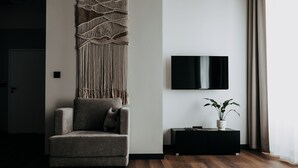 A modern living room with a grey sofa, a macrame wall hanging, a flat-screen TV, a black console table, and a potted plant.