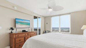 Quarto com cama, cômoda, ventilador de teto, televisão e janela com vista para o oceano.