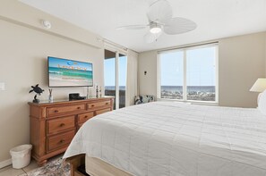 A bedroom with a bed, a dresser, a ceiling fan, a TV, and a window with a view of the ocean. - Gulf-front 2b - Island Princess - Free Beach Service and Paddleboard Rental! (Fort Walton Beach)