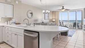 Uma cozinha com uma ilha grande, uma área de jantar com mesa e cadeiras, um ventilador de teto e vista para o oceano através de janelas amplas.