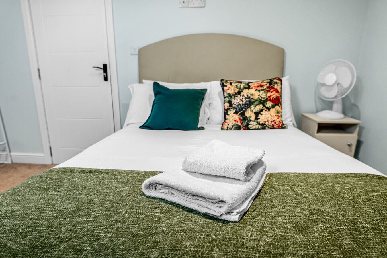 Un lit bien fait, recouvert d’une couverture verte, avec des oreillers blancs et un coussin à motifs floraux. Une serviette blanche pliée est posée sur le lit, et une table de chevet blanche se trouve à côté, avec un ventilateur dessus.