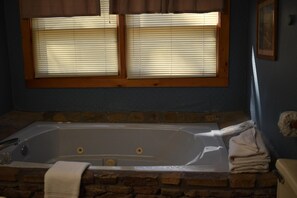 A bathroom with a stone wall, a bathtub, and two windows with blinds. - Family Traditions (Sevierville)