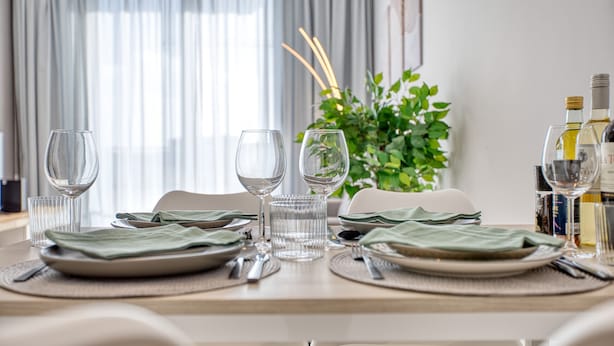 Uma mesa de jantar posta com pratos, talheres e copos, com uma planta e garrafas ao fundo.