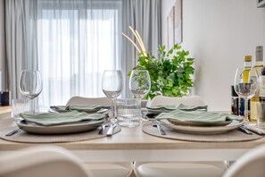 Uma mesa de jantar posta com pratos, talheres e copos, com uma planta e garrafas ao fundo.