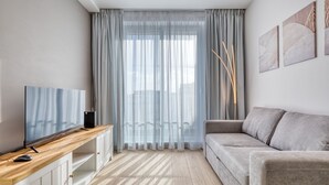 Uma sala de estar moderna com uma televisão de tela plana sobre um console de madeira, um sofá cinza e cortinas leves sobre uma janela grande.