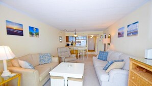 A living room with two sofas, a coffee table, a dining table with chairs, a kitchen area, and artwork on the walls. - Sandpiper Dunes 605 2 Bedroom Condo (Ocean City)