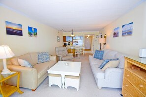 A living room with two sofas, a coffee table, a dining table with chairs, a kitchen area, and artwork on the walls. - Sandpiper Dunes 605 2 Bedroom Condo (Ocean City)