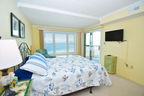 A bedroom with a bed, a chair, a TV, a window with a view of the sea, and a balcony. - Sandpiper Dunes 605 2 Bedroom Condo (Ocean City)