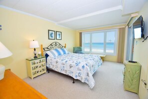 A bedroom with a bed, bedside table, lamp, chair, and a TV. - Sandpiper Dunes 605 2 Bedroom Condo (Ocean City)
