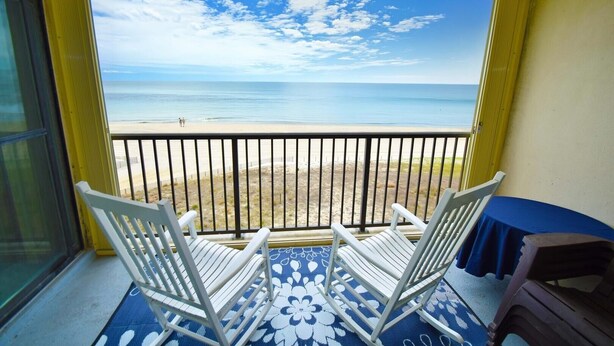 A balcony with a view of the beach, white chairs, and a blue table. - Sandpiper Dunes 605 2 Bedroom Condo (Ocean City)