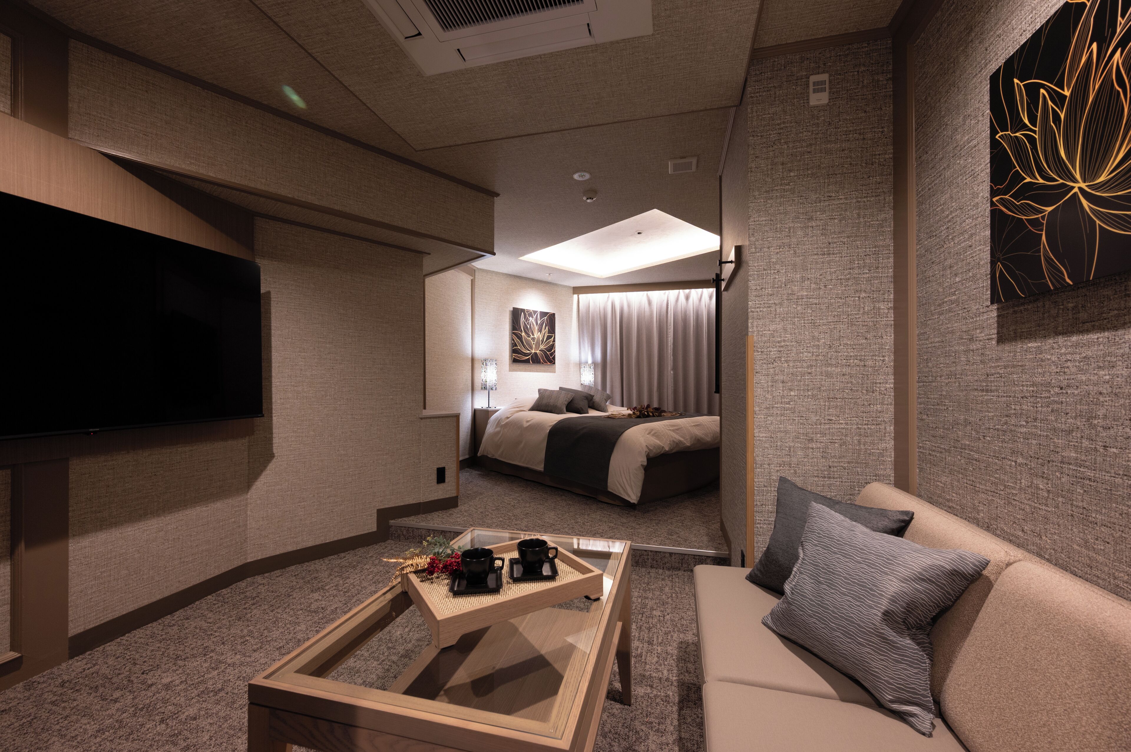 A modern hotel room with a flat-screen TV, a sofa, a coffee table with two cups, and a bed with a view of the city.