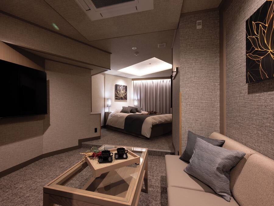 A modern hotel room with a flat-screen TV, a sofa, a coffee table with two cups, and a bed with a view of the city.