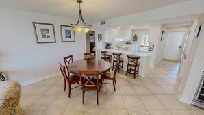 A spacious dining area with a round wooden table and chairs, a kitchen with white cabinets, and a large window providing natural light. - Mainsail 136 3 Bedroom Condo (Miramar Beach)