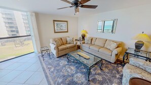 A living room with a sofa set, a glass coffee table, a ceiling fan, and a view of the ocean through a large window. - Mainsail 136 3 Bedroom Condo (Miramar Beach)