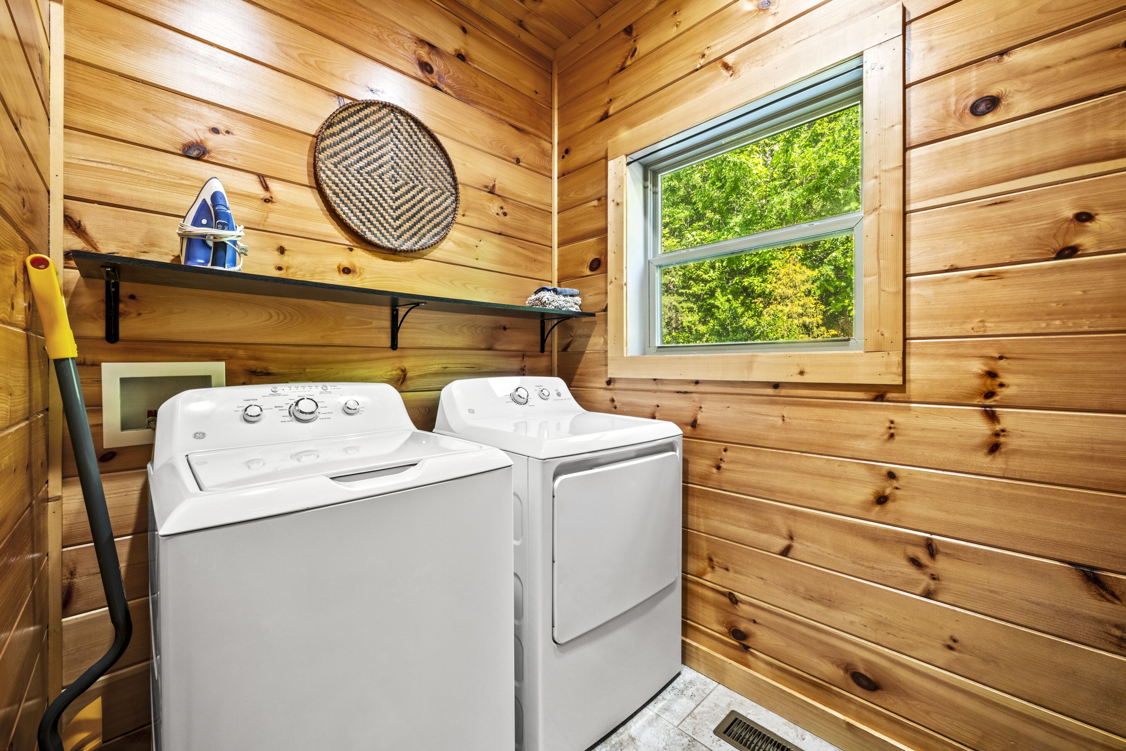 A laundry room with two white washing machines, a wooden wall, a window with a view of greenery, and a shelf with a woven basket and a small blue object.