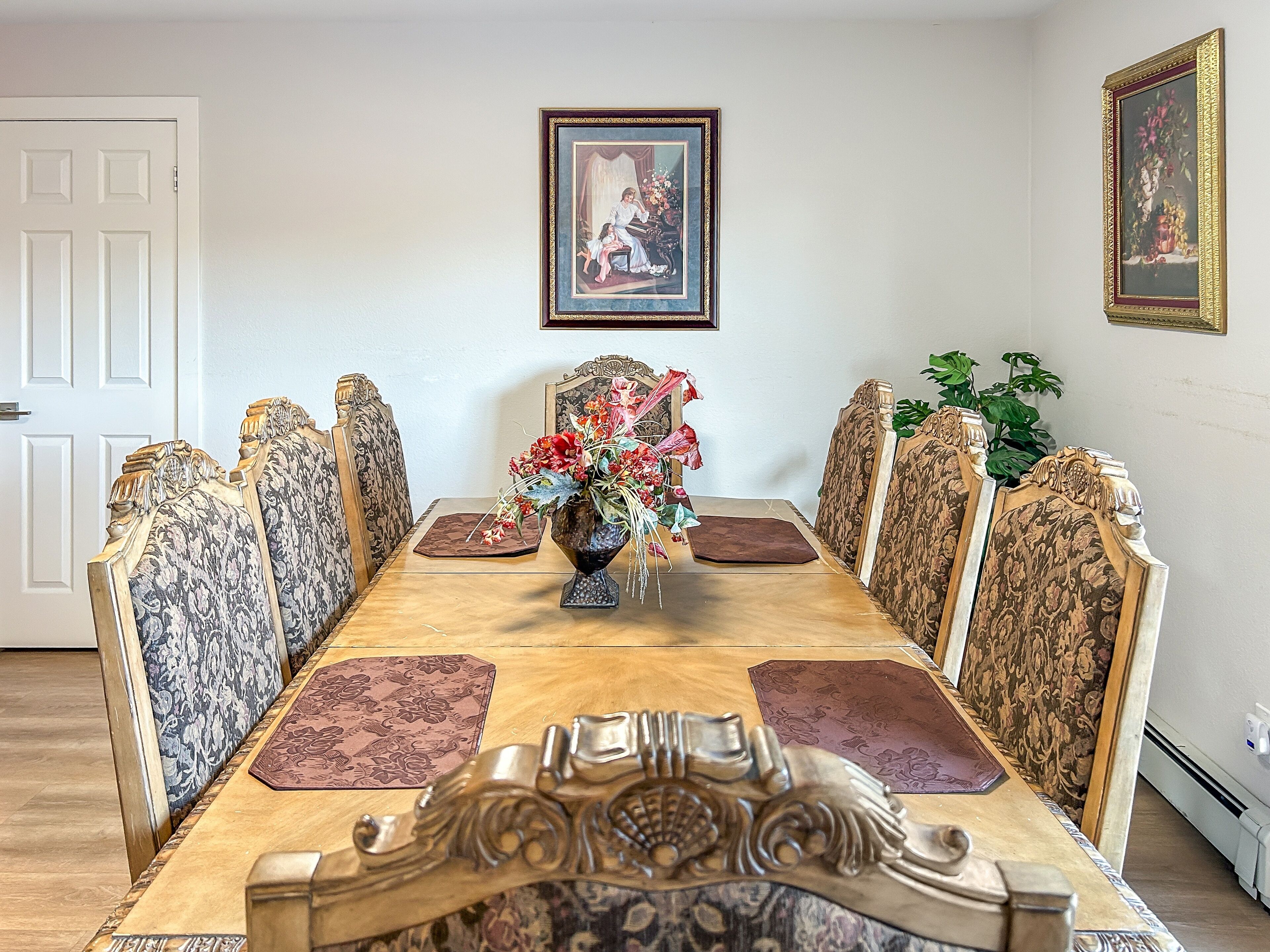 A dining room with a long wooden table, ornate chairs, a vase of flowers, and framed pictures on the walls.