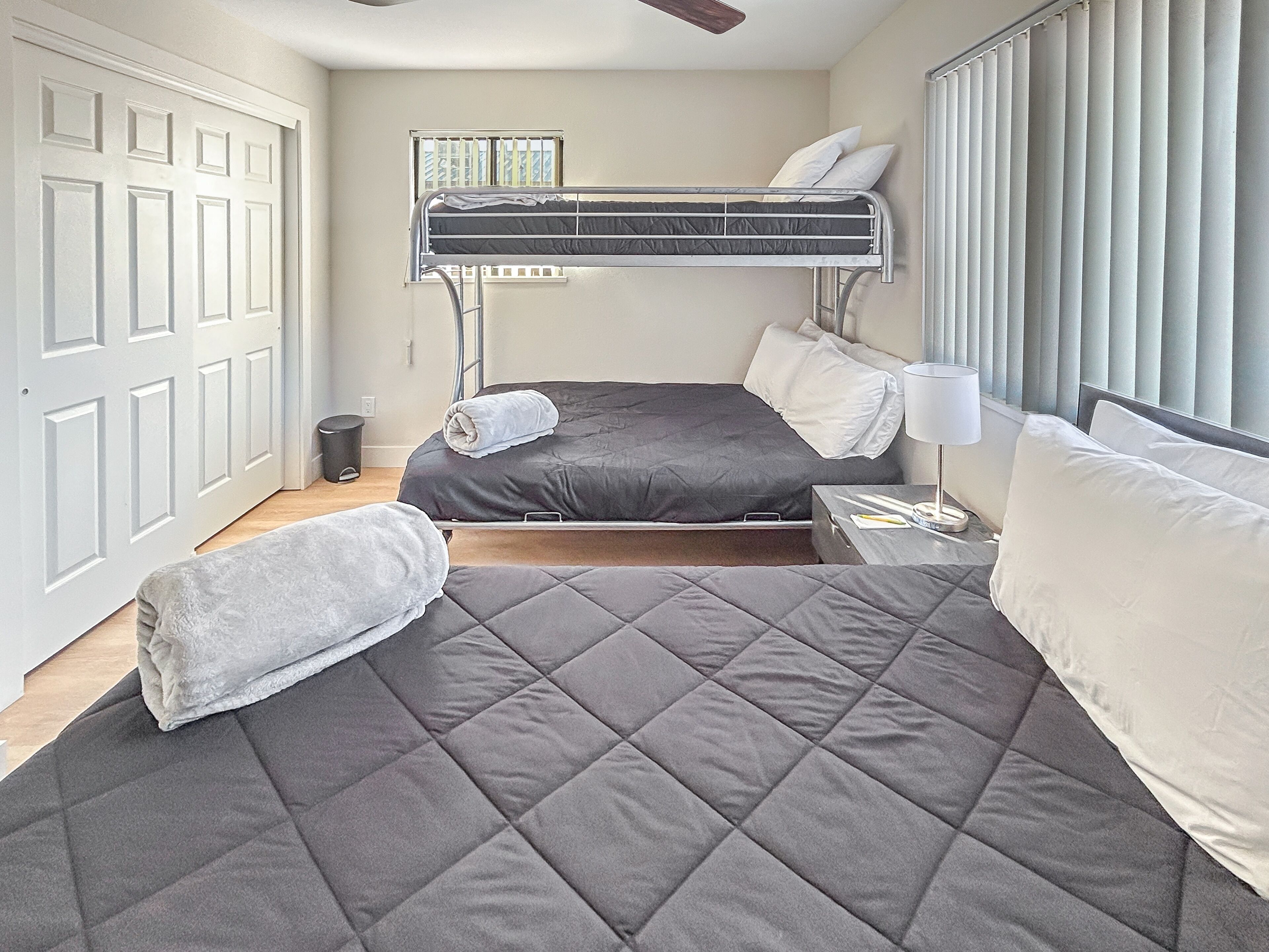 Quarto com cama de solteiro, uma cama individual, mesa de cabeceira com abajur e janela com persianas.