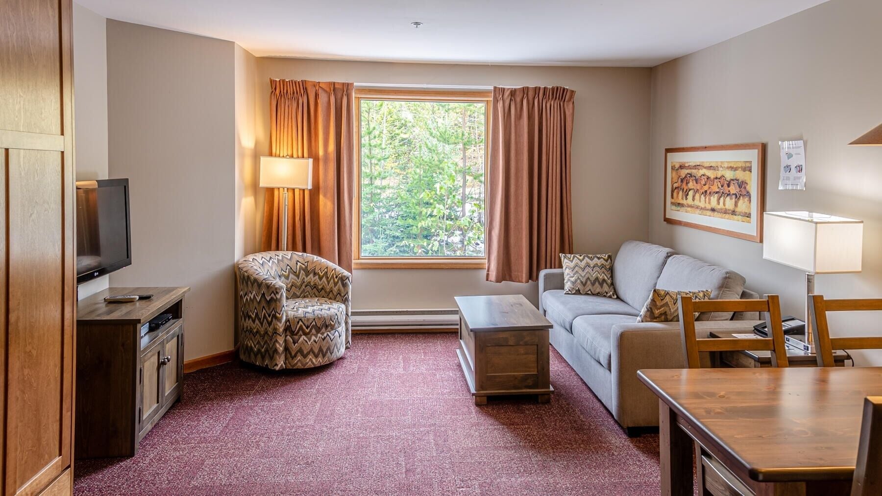 Habitación de hotel compacta con televisión, sofá, sillón, una mesita y ventana con cortinas.