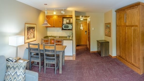 Cocina compacta con armarios de madera, microondas y un lavabo. Hay una zona de comedor con mesa y sillas. El suelo está cubierto con una alfombra estampada.