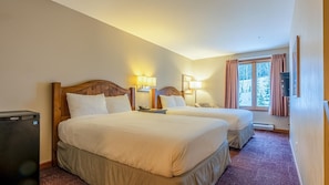 A hotel room with two beds, a wooden headboard, bedside lamps, a heater, and a window with curtains. - Cahilty #225 Hotel Room (Sun Peaks)