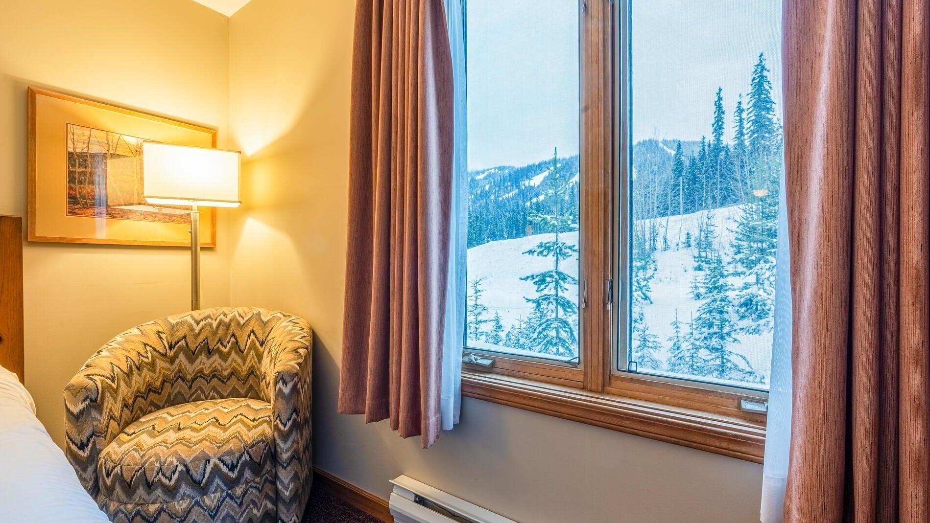A cozy room with a large window offering a snowy mountain view, a patterned armchair, and a framed picture on the wall.