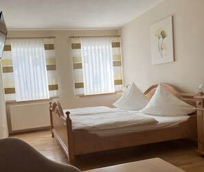 Une chambre à coucher avec un lit en bois, deux fenêtres avec des rideaux, un radiateur et une lampe fixée au mur.