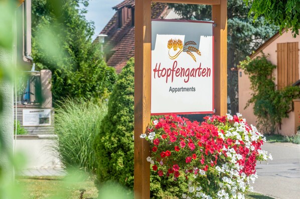 Letrero de Hopfengarten Apartments con un logotipo de uvas y hojas, rodeado de un macizo de flores rojas y blancas.