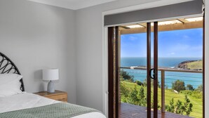 A bedroom with a bed, a bedside table with a lamp, and a view of the ocean through a sliding glass door. - Ohana Oasis, Kiama, South Coast (Kiama)