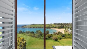 A coastal view from a balcony with a clear sky, a beach, and a residential area in the background. - Ohana Oasis, Kiama, South Coast (Kiama)