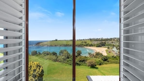 A coastal view from a balcony with a clear sky, a beach, and a residential area in the background. - Ohana Oasis, Kiama, South Coast (Kiama)