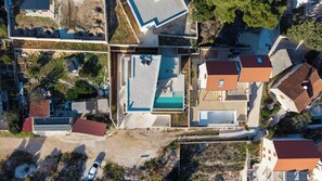 An aerial view of a residential area with a swimming pool, multiple houses with red roofs, and a mix of paved and unpaved surfaces. - Villa Altamare (Splitska)