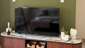 Un televisor de pantalla plana sobre un mueble de madera con encimera de mármol, un aire acondicionado montado en la pared, y un cuadro enmarcado encima del televisor.