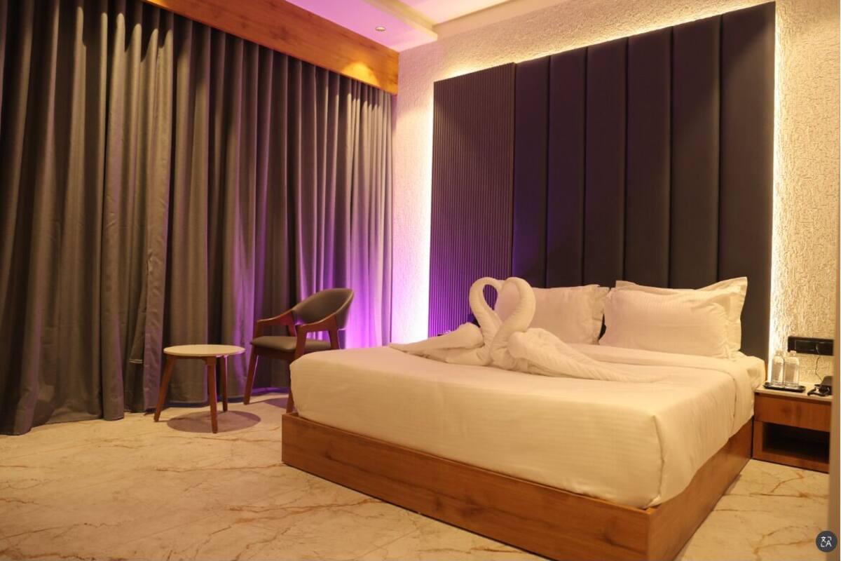 A hotel room with a large bed, a wooden headboard, a small round table, a chair, and a nightstand with a lamp.