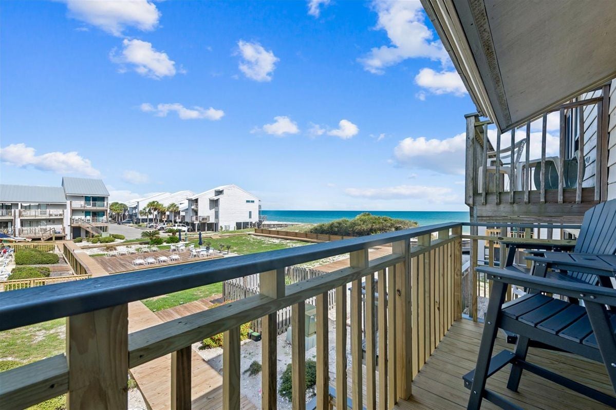 A balcony with a view of the beach and ocean, wooden deck, and outdoor seating.