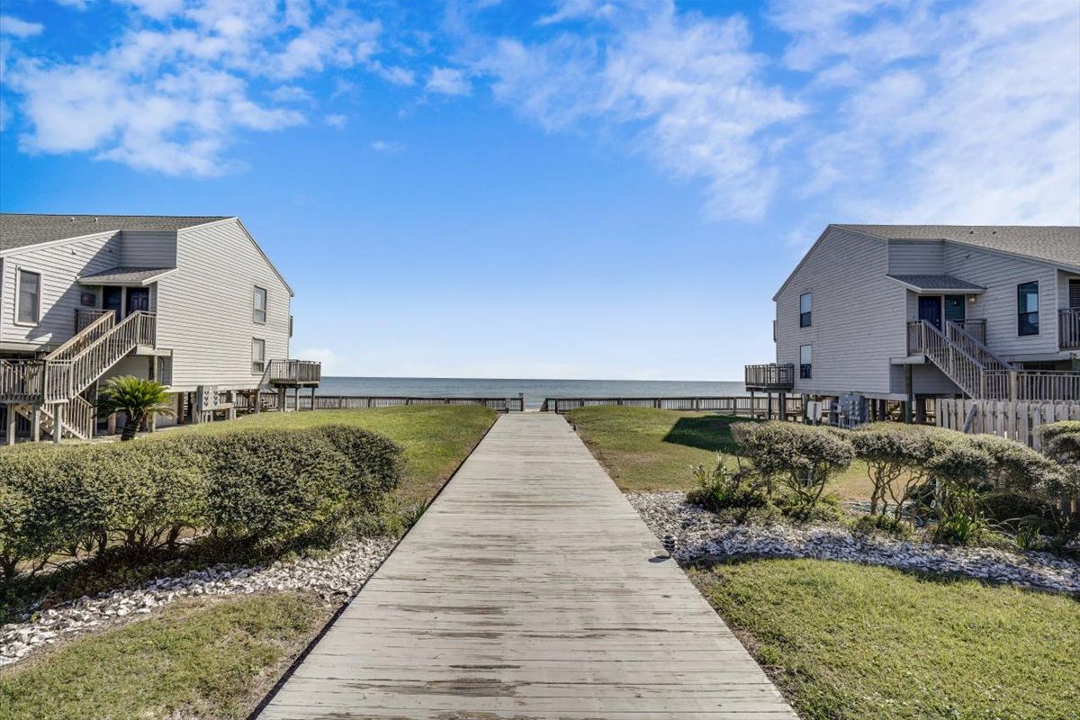 A wooden boardwalk leading to a beachfront property with two-story buildings, a clear sky, and a view of the ocean.
