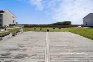 Eine Holzstege führt zu einem Strand mit einer Rasenfläche und einem Gebäude auf der linken Seite.