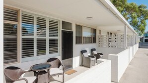 A covered outdoor seating area with wicker chairs and a glass table, adjacent to a building with large windows and a white exterior. - Palm Beach Villas (Rockingham)