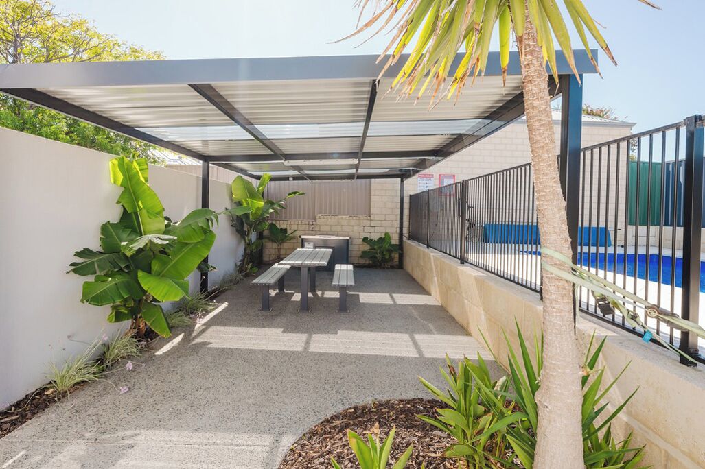 A covered patio area with a pool, featuring a table and benches, surrounded by plants and a palm tree.