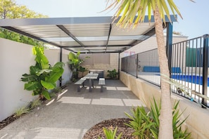 Un patio cubierto con piscina, que incluye una mesa y bancos, rodeado de plantas y una palmera.