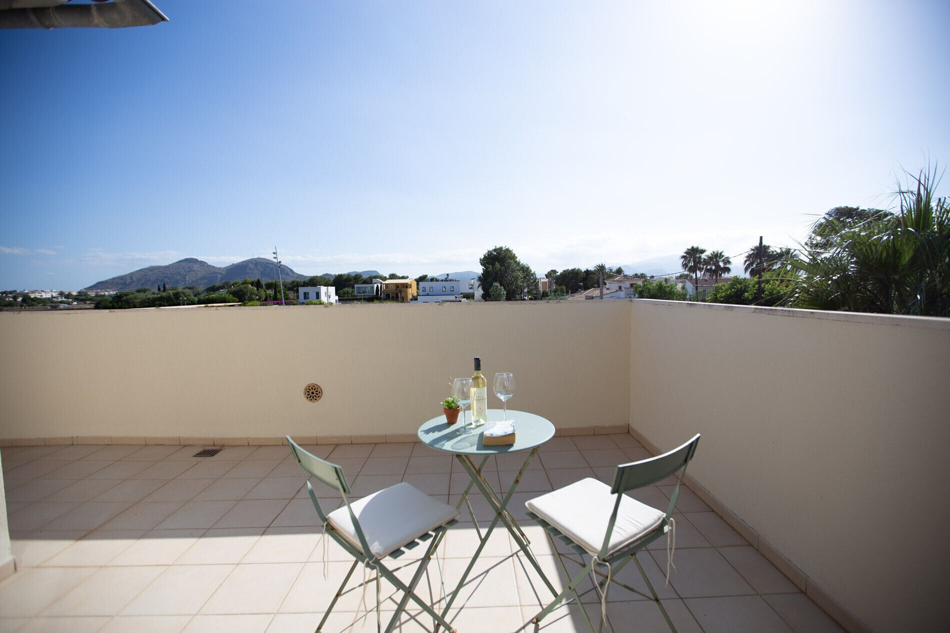 Une petite table ronde, accompagnée de deux chaises, est placée sur une terrasse carrelée, d’où l’on a vue sur un quartier résidentiel avec ses maisons et ses arbres.