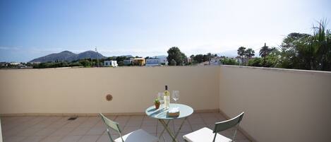 Une petite table ronde, accompagnée de deux chaises, est placée sur une terrasse carrelée, d’où l’on a vue sur un quartier résidentiel avec ses maisons et ses arbres.