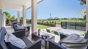 Un área de estar techada al aire libre con muebles de mimbre, una mesa con una bandeja de tazas y platos, y vistas a una piscina y un paisaje lejano.