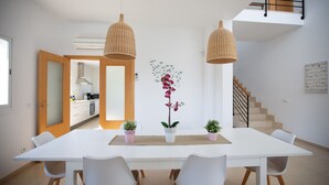 Un comedor moderno con una mesa blanca, cuatro sillas, dos lámparas de mimbre colgantes y una orquídea en maceta como centro de mesa.