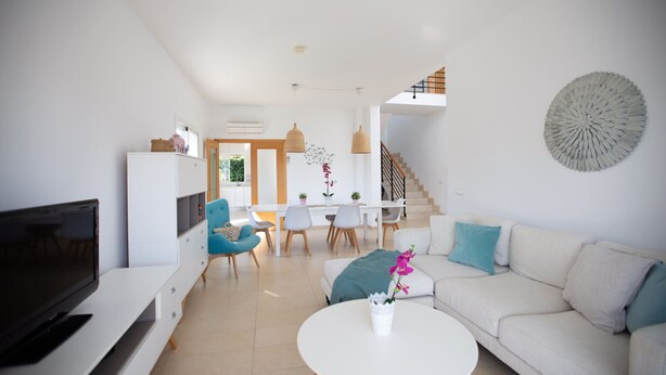 Una sala moderna con un sofá blanco, una mesa de centro y un televisor. Hay un comedor con una mesa y sillas, y una escalera que lleva a otro nivel.
