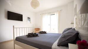Un dormitorio con cama, televisor de pantalla plana en la pared, ventana con cortinas y una silla.