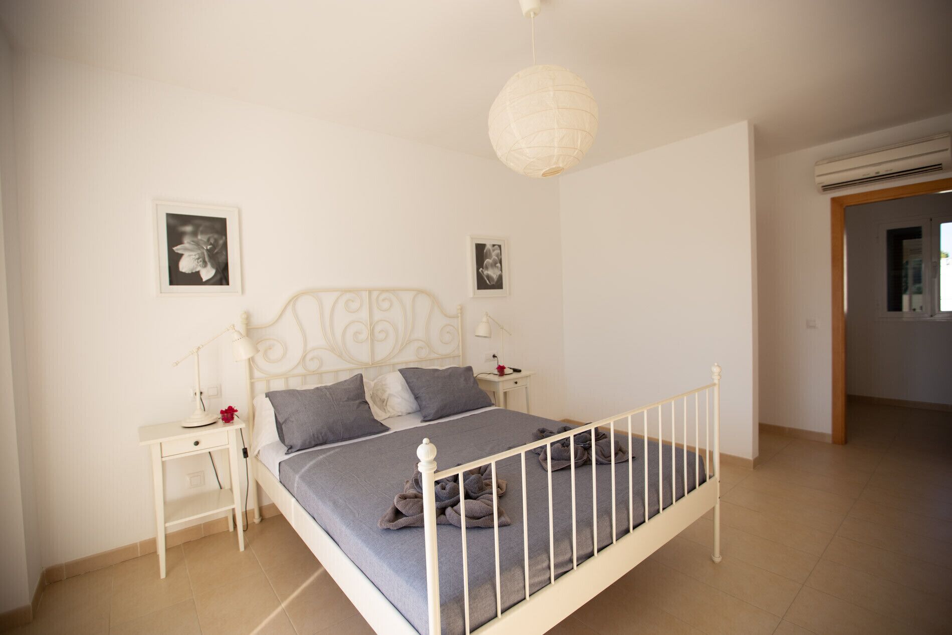 A bedroom with a white metal bed frame, a grey bedspread, two bedside tables with lamps, and a white ceiling light.