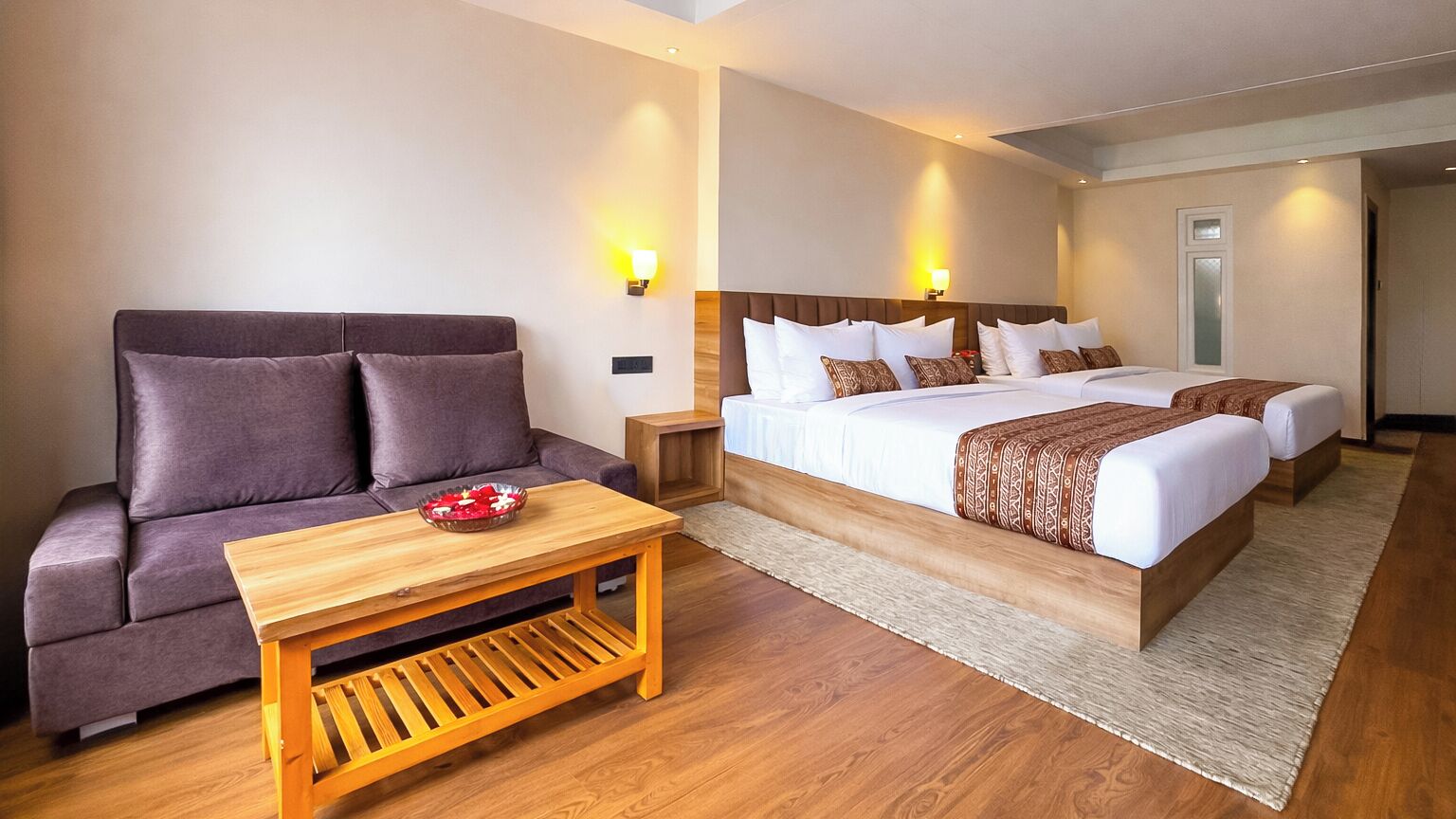 A hotel room with a sofa, a wooden coffee table, and a double bed with a patterned bedspread.